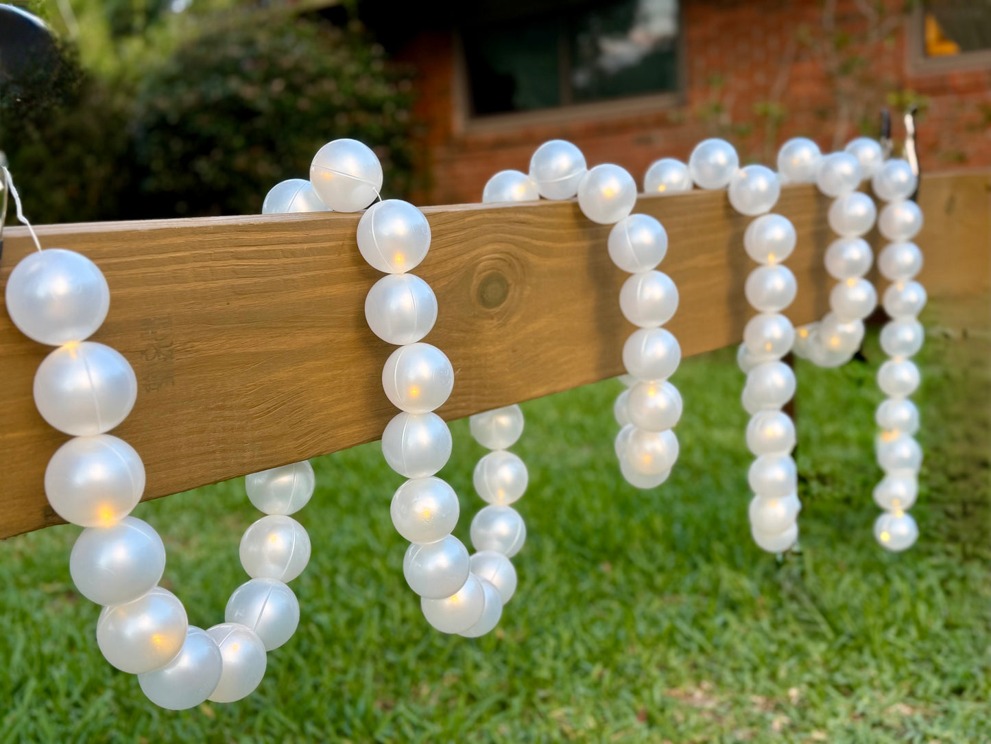 White faux-pearl bead garlands with warm white string lights draped over a wooden fence on a green lawn.
