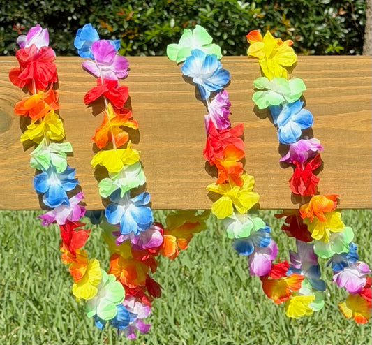 Four colorful Hawaiian-style flower leis garland draped over a wooden railing outdoors, with green grass and foliage in the background.