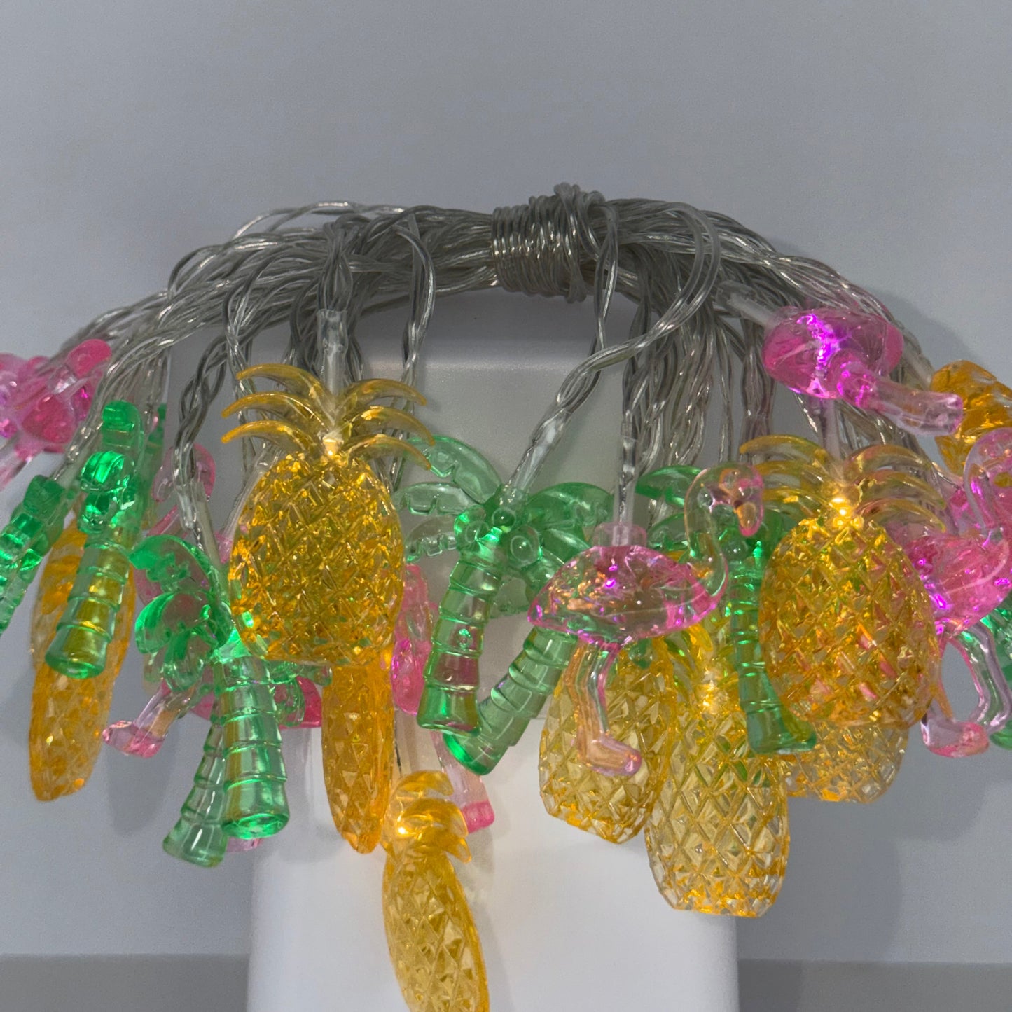 Light-up plastic pineapple string lights with yellow, green, and pink glowing bulbs on a silver wire, shown on a white display stand against a light gray background.