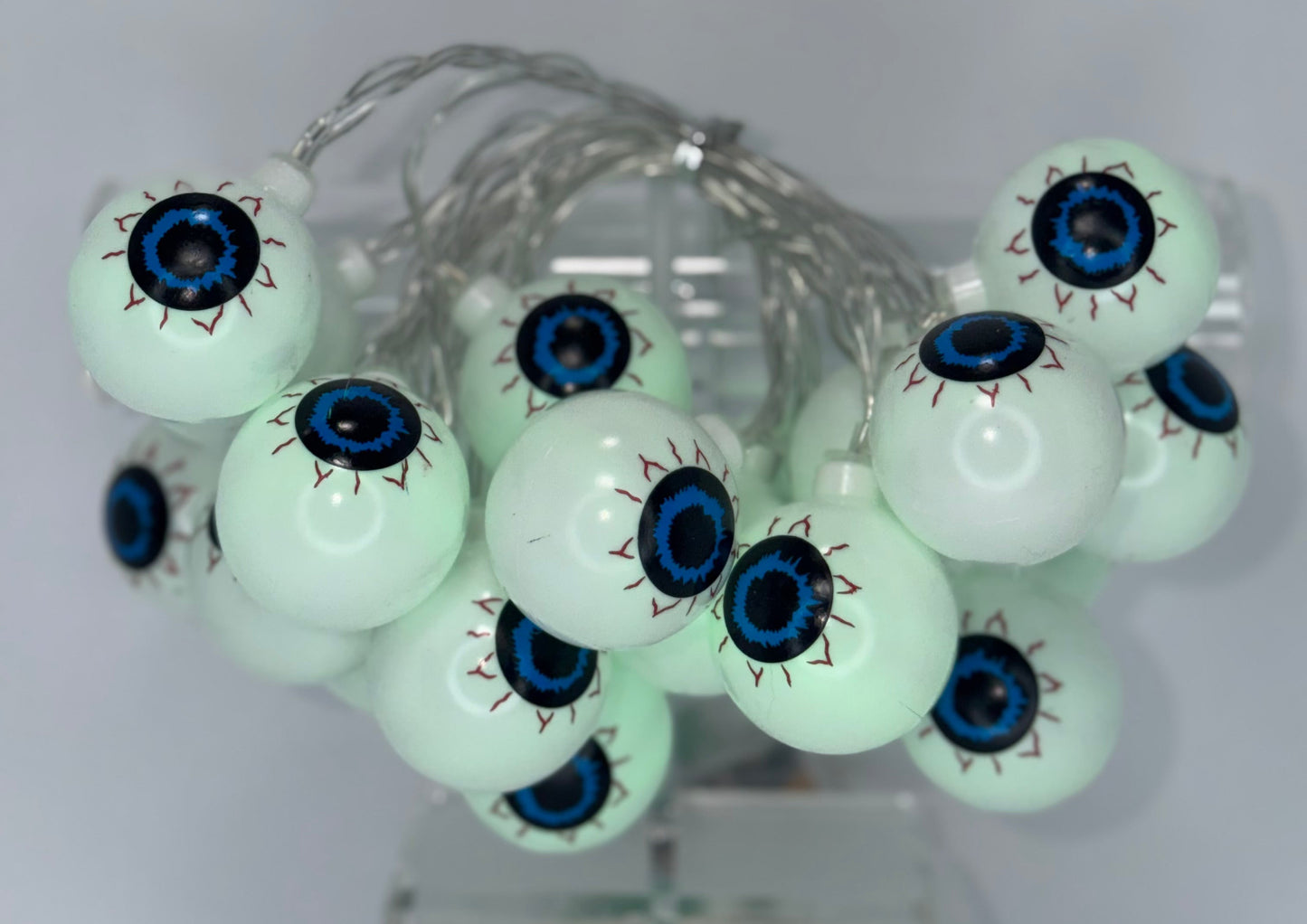 Glowing pale green bony eyeball string lights with realistic blue and black pupils, arranged on a clear stand against a neutral gray background.