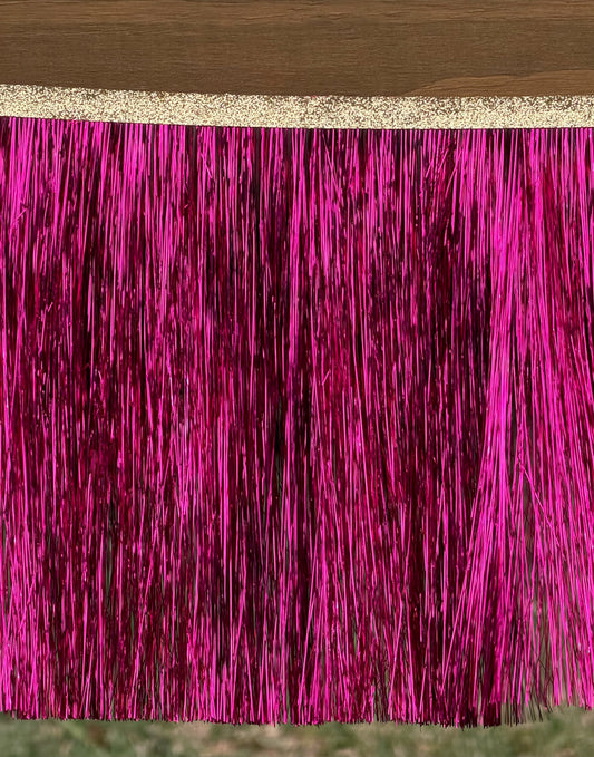 Bright hot pink metallic fringe trim hanging from a glittery gold ribbon, shown against a dark wood surface with a hint of grass below.