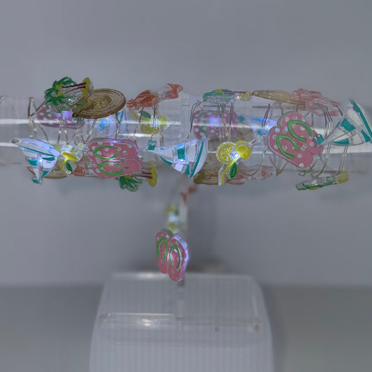 A clear cylindrical display tube mounted on a white plastic stand, filled with colorful iridescent foil sheets and small decorative elements, set against a plain light gray background.