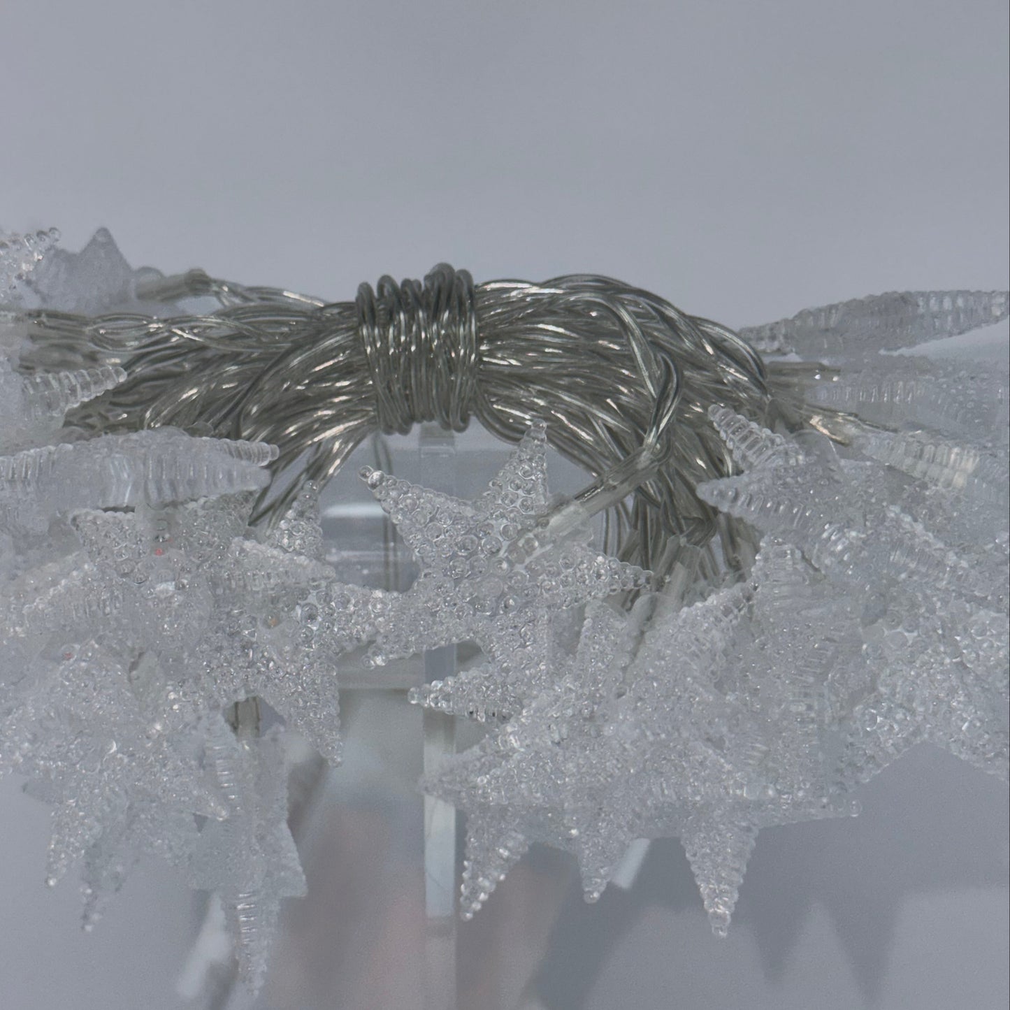A close-up of a decorative silver wire wreath or garland wrapped with textured clear plastic mesh, shown against a pale gray background.