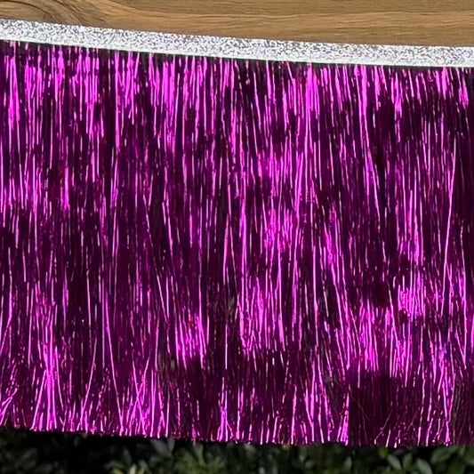 A close-up of a metallic tinsel fringe curtain in bright magenta-purple, with a silver glittery top edge, photographed against a wood surface and dark green background.