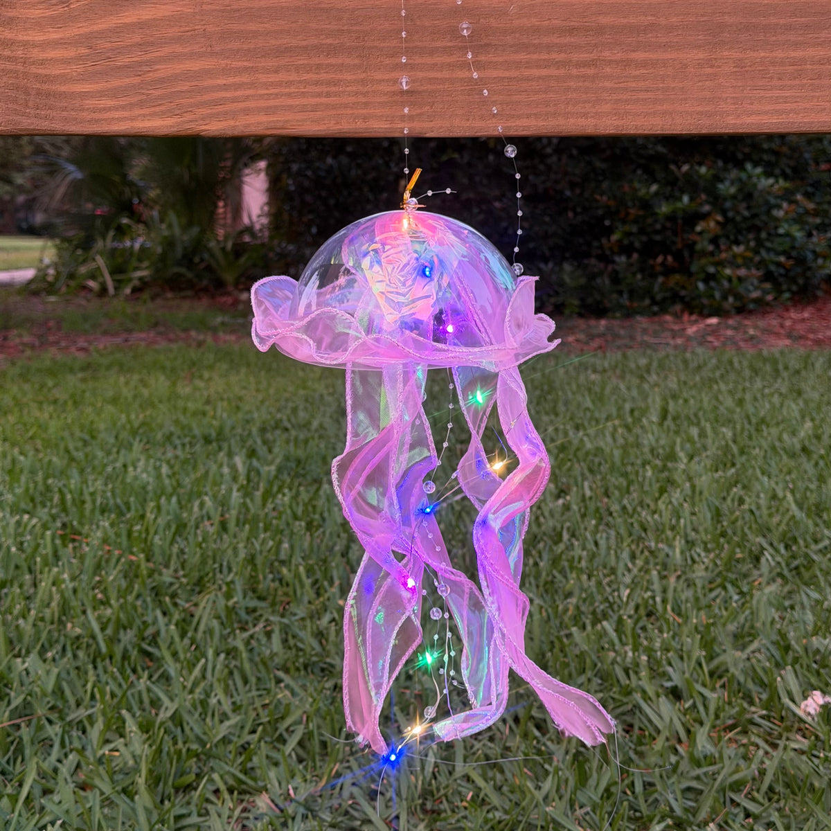 A jellyfish-shaped outdoor hanging decoration made of sheer pink mesh with embedded multicolor LED lights, suspended over a grassy lawn under a wooden fence.