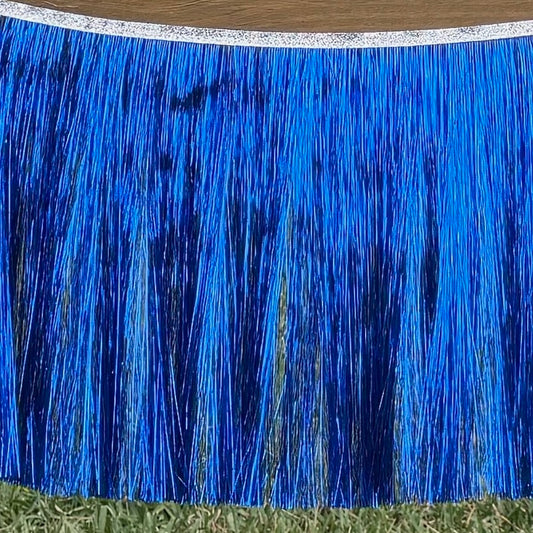A bright blue metallic fringe banner hanging outdoors on grass, with a silver glittery top band and a wood surface visible above it.