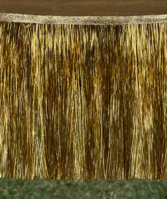 A close-up of a gold metallic fringe trim hanging from a wooden rod, with a textured gold band at the top and strands extending down to a blurred green ground background.