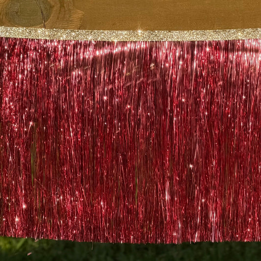 A close-up of a red metallic tinsel fringe curtain with a glittery gold top band, shown hanging against a wooden surface.