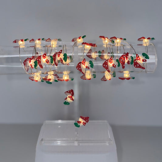 A clear acrylic holiday light display stand holding multiple small LED Christmas lights with warm white bulbs and red, green, and gold ornaments, mounted on a white plastic base against a plain gray studio background.