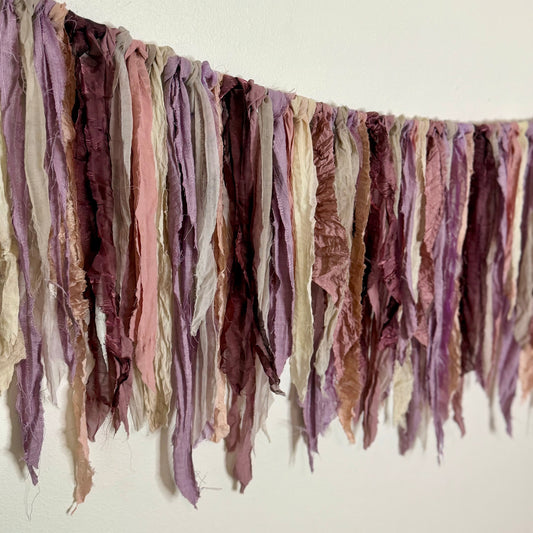 A rustic decorative garland made of hanging distressed fabric strips in muted dusty purple, mauve, blush pink, taupe, and cream, strung across a plain off-white wall.