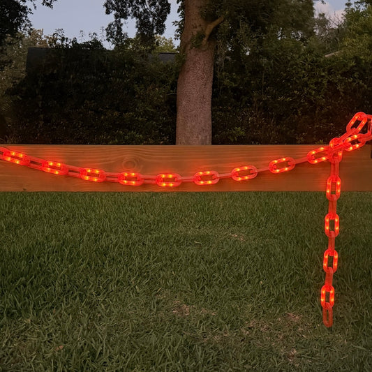 A red illuminated safety chain barrier draped across a wooden fence in an outdoor backyard setting with green grass and trees in the background.