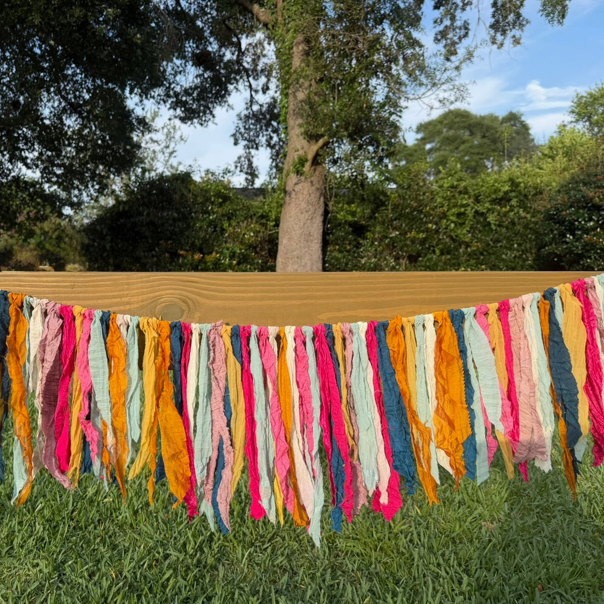 A colorful fabric fringe garland hanging outdoors on a wooden fence over green grass, featuring vertical strips of pink, orange, yellow, blue, and white.