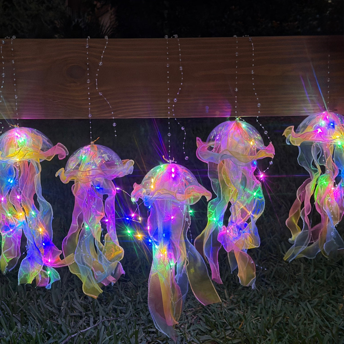 Glowing jellyfish garden lights hanging from a wooden fence on a grassy lawn at dusk, with iridescent translucent bodies and small string lights.