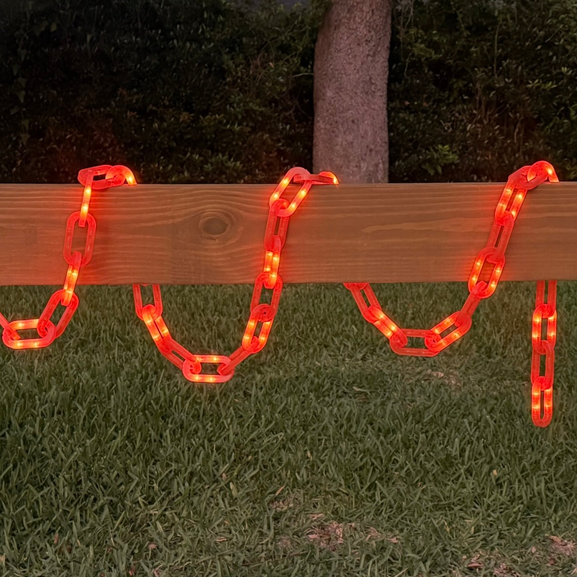 Red glowing chain-link outdoor lights draped over a wooden fence on a grassy lawn at night.