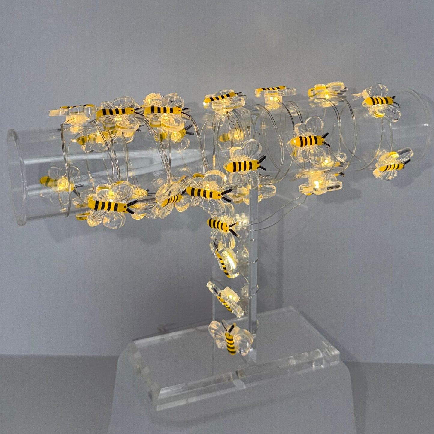 A modern wall-mounted light fixture with a clear acrylic cylindrical body decorated with numerous small yellow and black bumblebee figures, casting a warm glow against a plain light gray wall.
