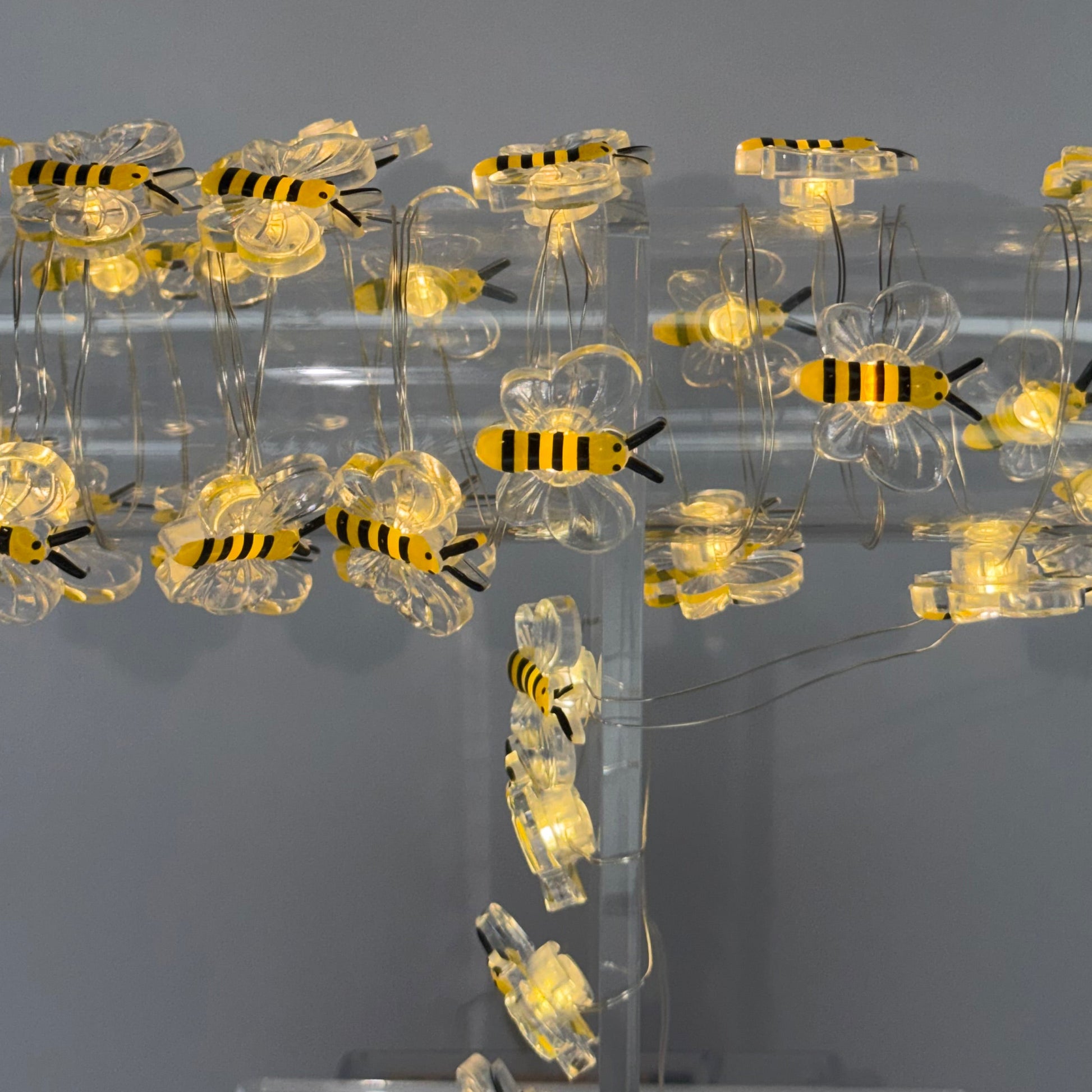 A string of decorative bumblebee LED fairy lights glowing with warm yellow light, attached to a clear acrylic branch-like structure against a plain gray background.