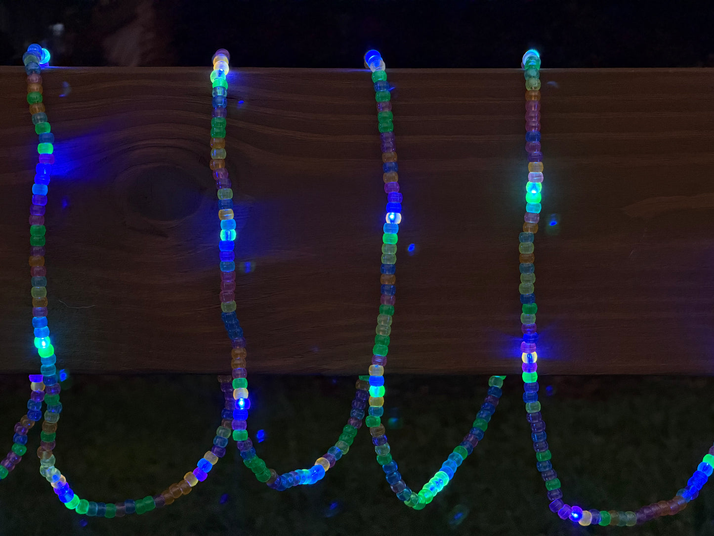 Multicolor LED beaded garland draped over a wooden fence outdoors at night, glowing with blue, green, and purple lights.