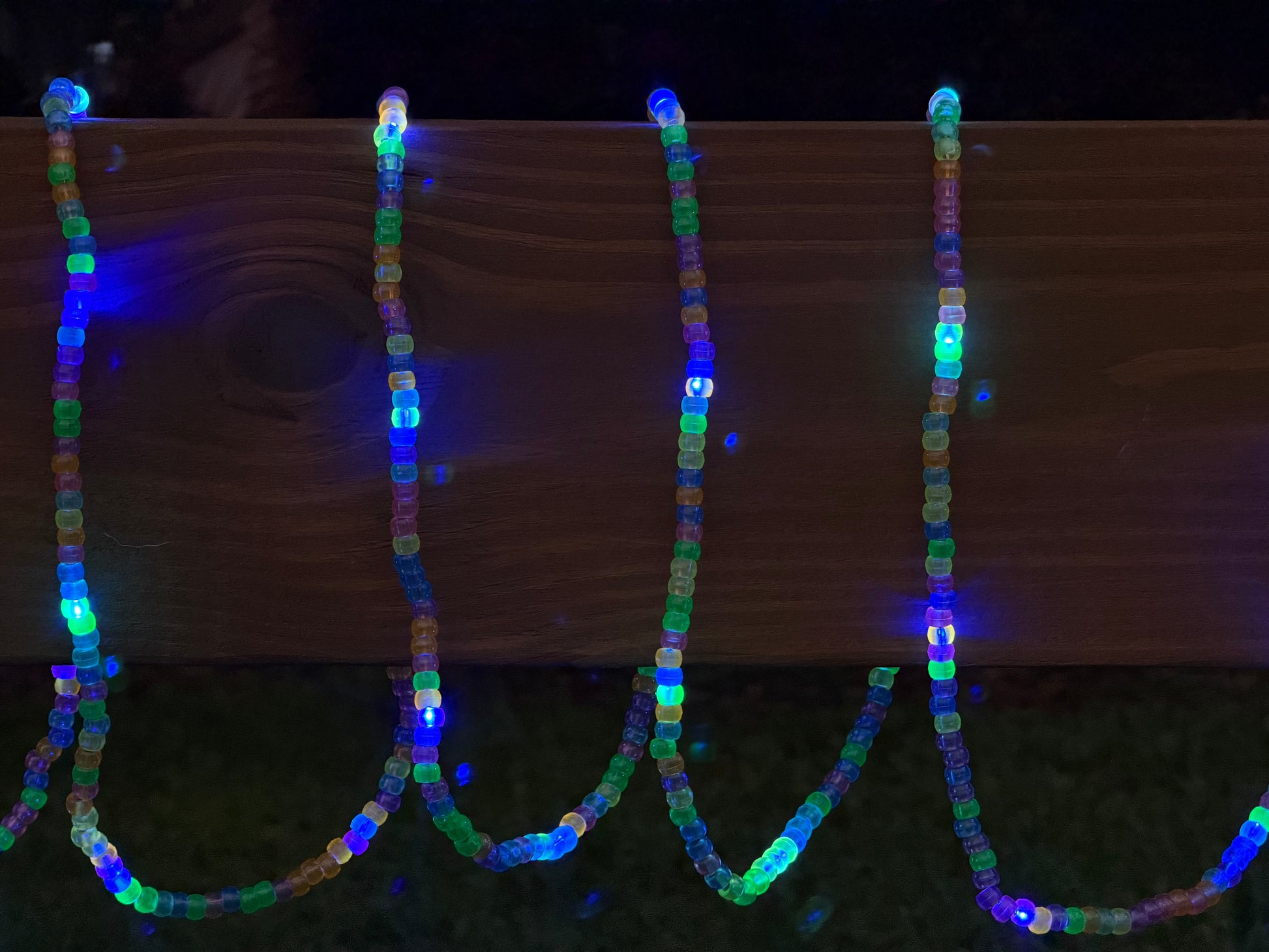Multicolor LED beaded garland draped over a wooden fence outdoors at night, glowing with blue, green, and purple lights.