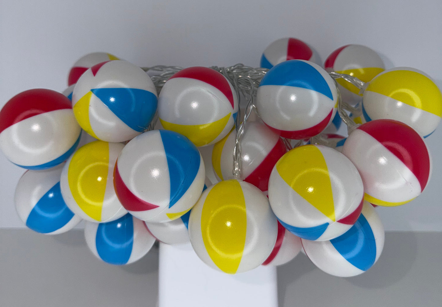 A cluster of miniature beach ball string lights with red, white, blue, and yellow striped spheres on a silver wire, shown hanging against a plain light gray wall.