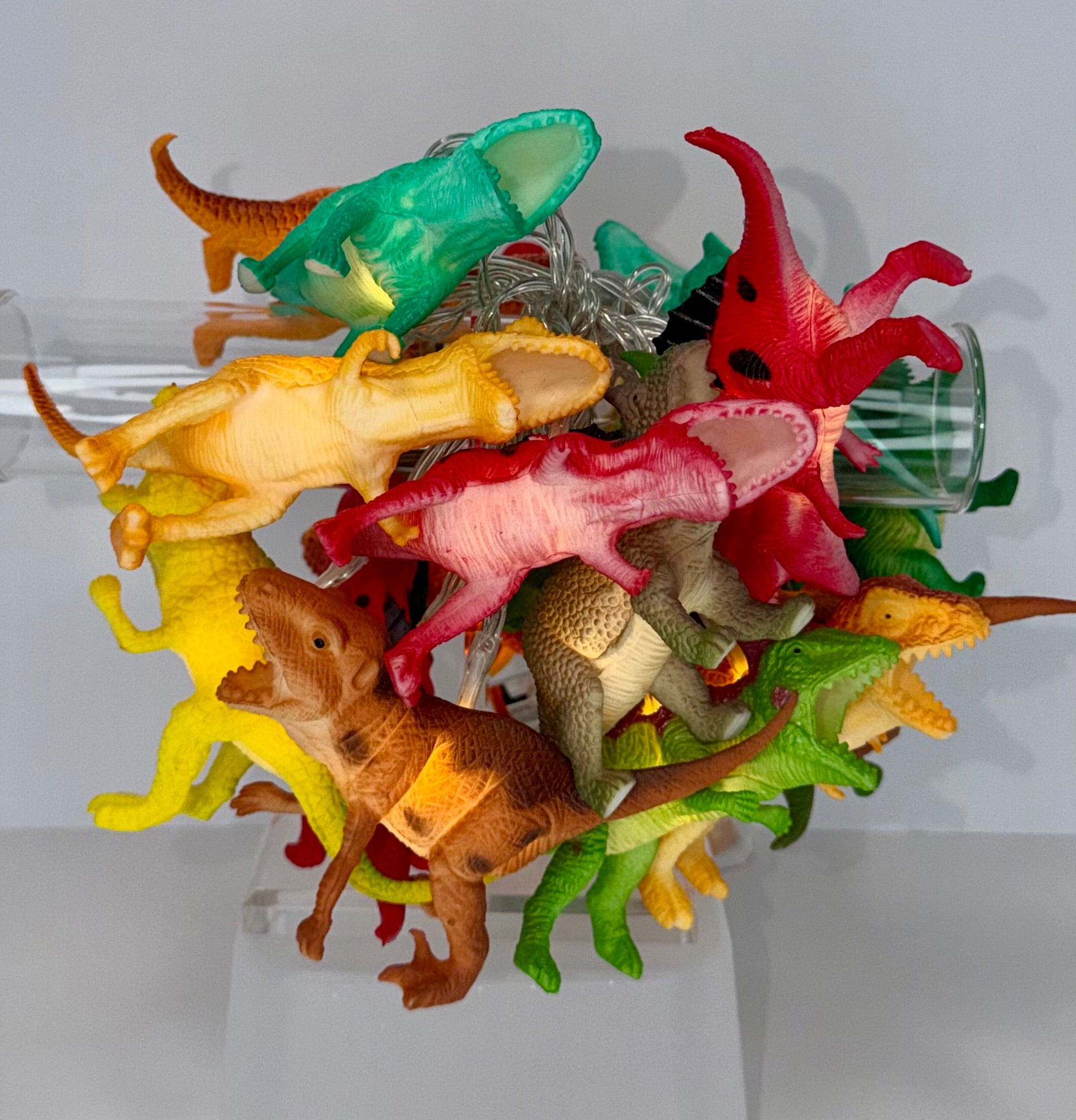 A collection of colorful plastic toy dinosaurs in various species and colors, arranged on a clear plastic display stand against a light gray background.