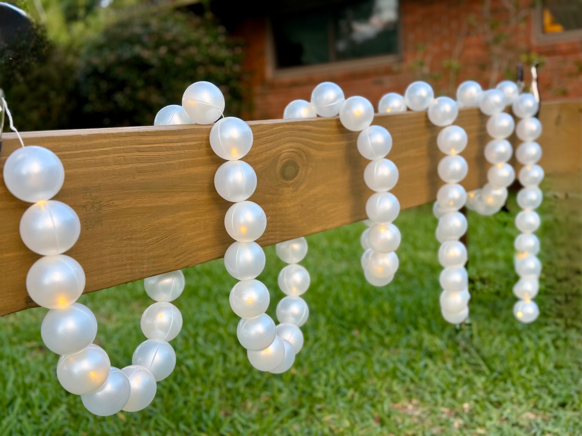 White faux-pearl bead garlands with warm white string lights draped over a wooden fence on a green lawn.
