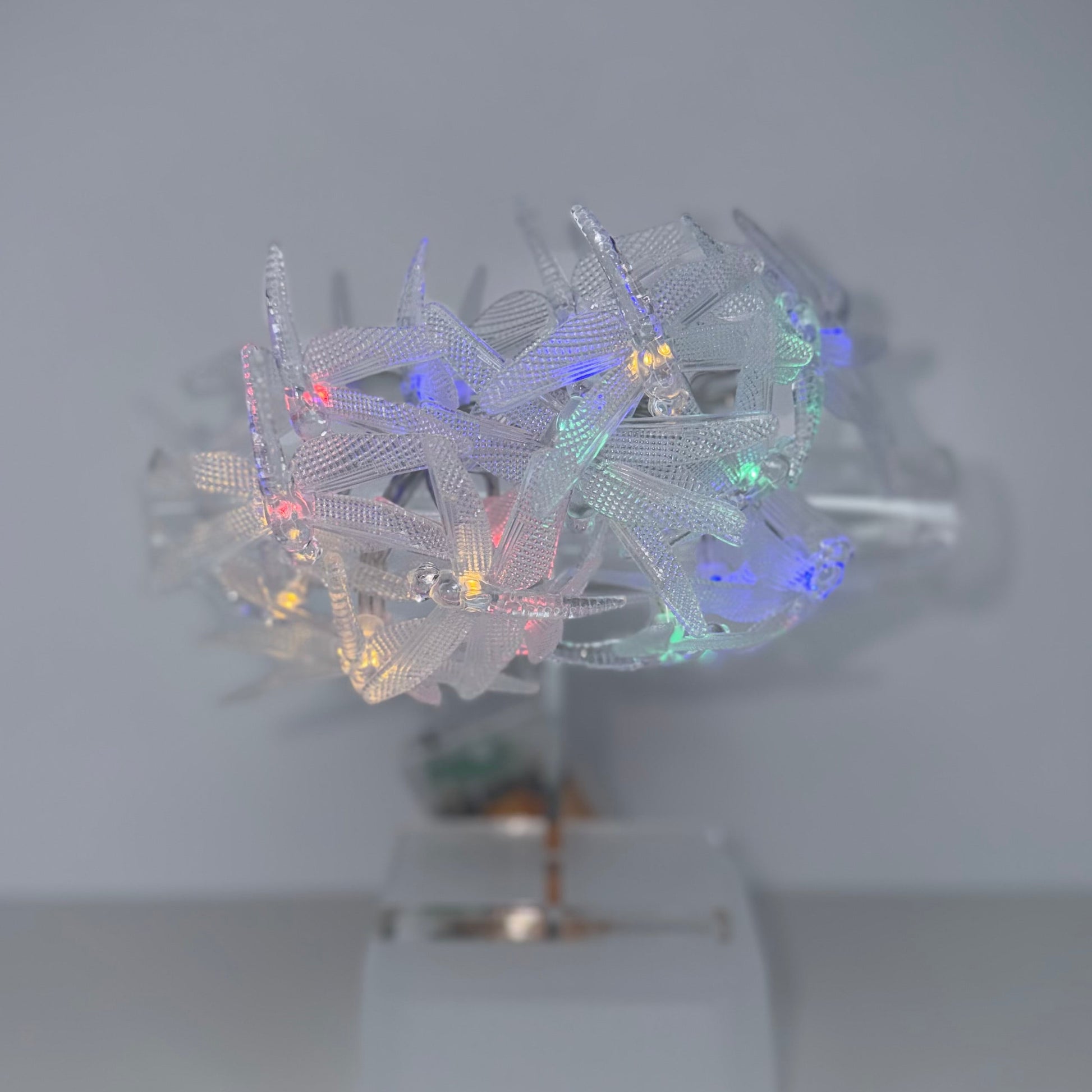 A white sheer mesh headband with small multicolored LED fairy lights, displayed on a clear stand against a plain light gray background.