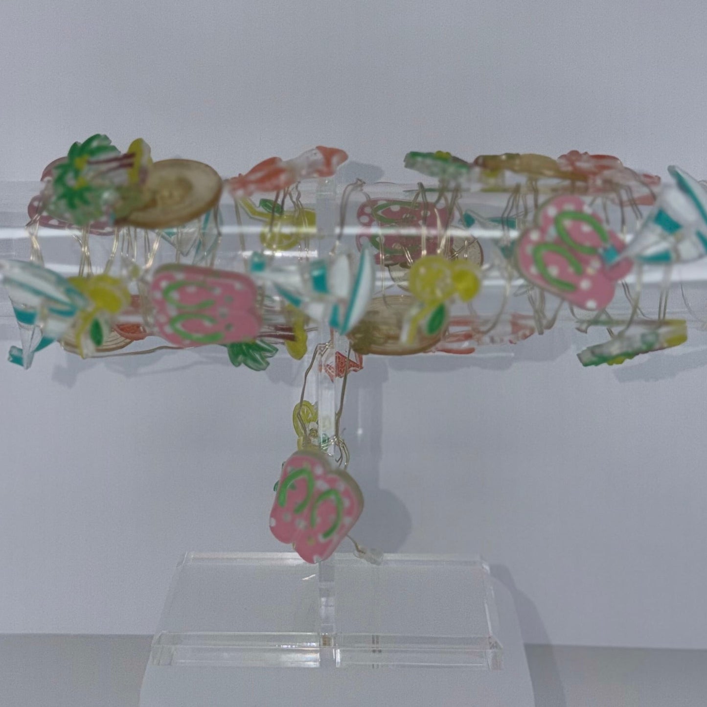 A clear acrylic display stand holding a cylindrical transparent object covered with small multicolored floral or decorative elements, shown against a plain light gray background with a white power cord visible at the lower right.