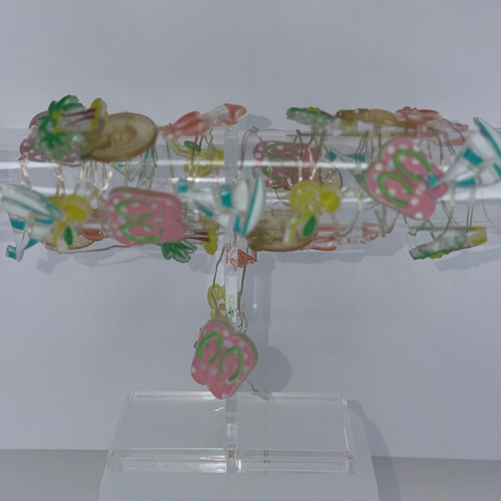 A clear acrylic display stand holding a cylindrical transparent object covered with small multicolored floral or decorative elements, shown against a plain light gray background with a white power cord visible at the lower right.