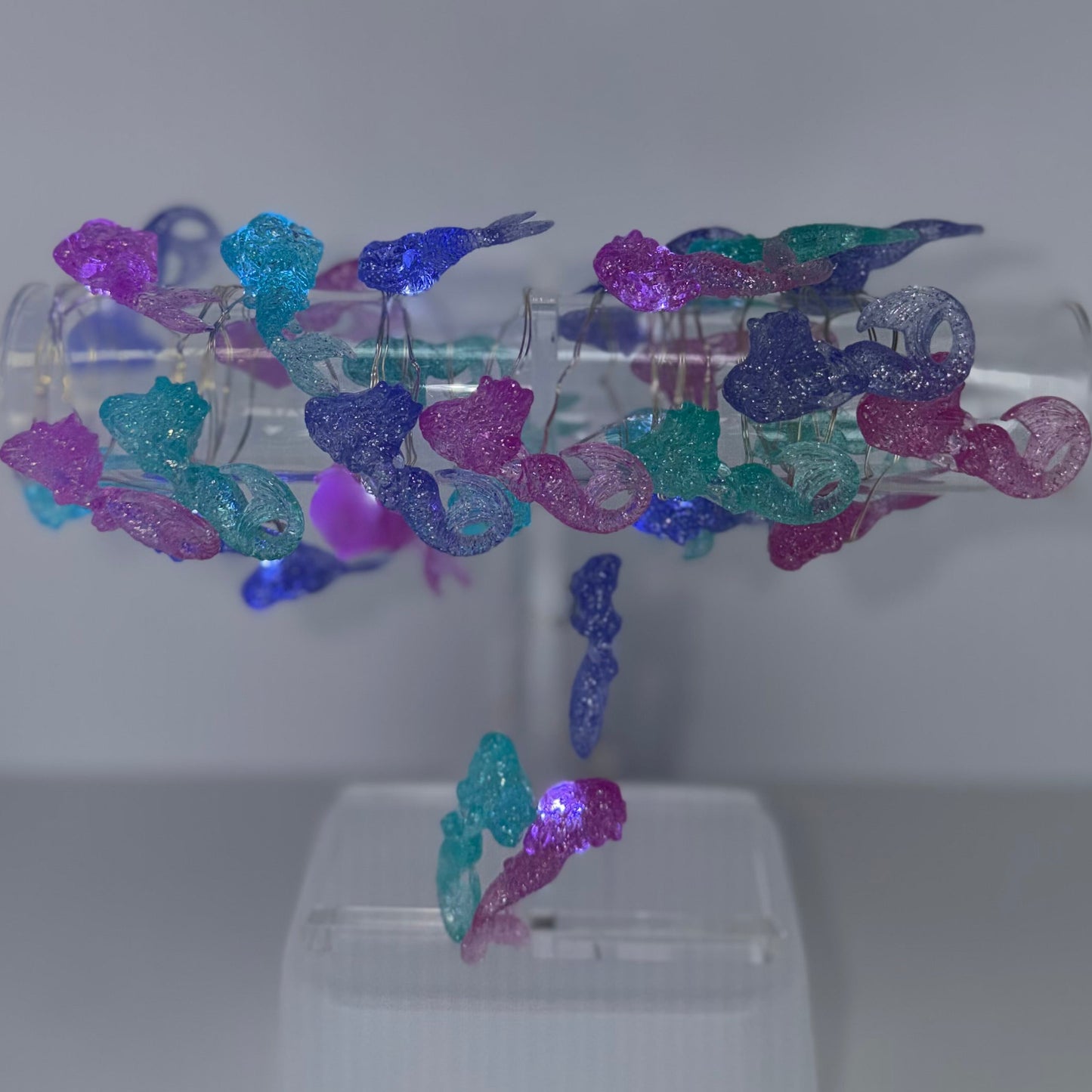 A clear acrylic jewelry display stand holding a ring with multiple dangling clear and iridescent butterfly-shaped charms in pink, purple, blue, and teal, set against a plain light gray background.