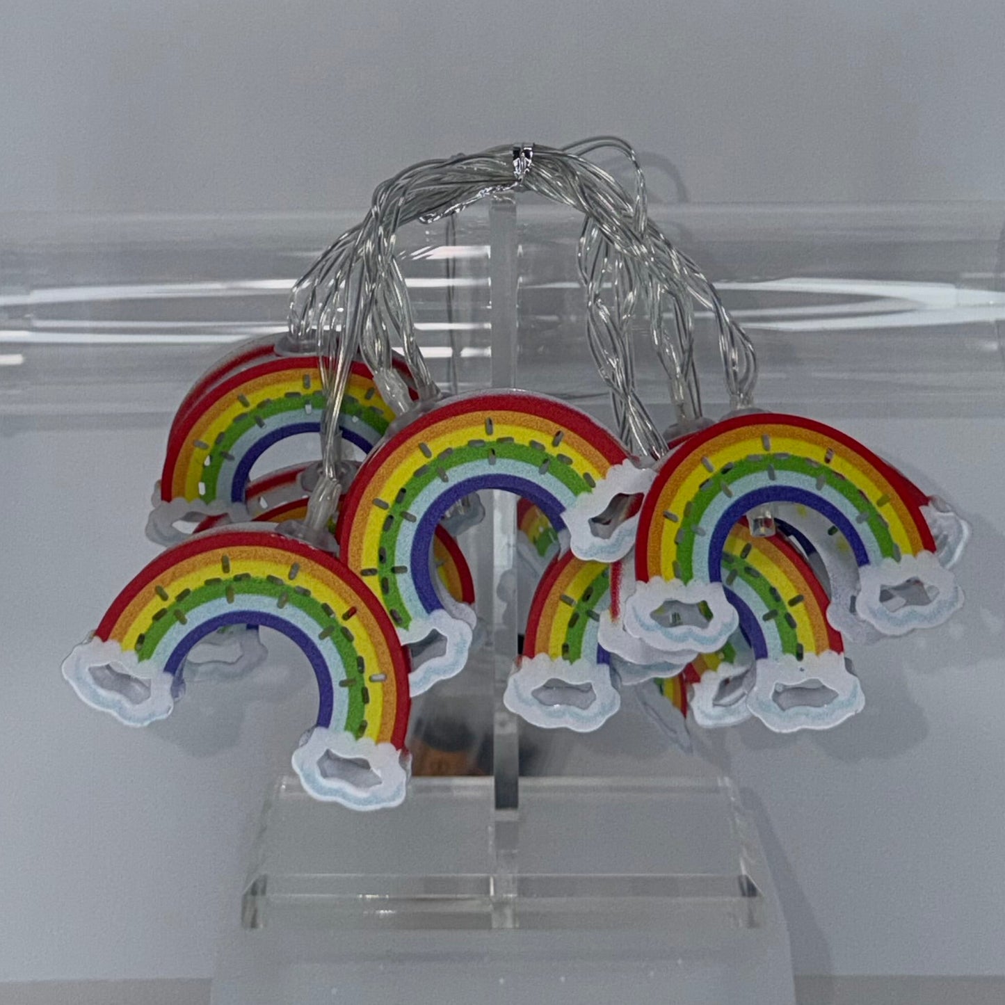 A set of small rainbow-shaped fairy lights with white cloud ends and a silver wire, displayed hanging from a clear acrylic stand in front of a transparent cylindrical tube against a light gray background.