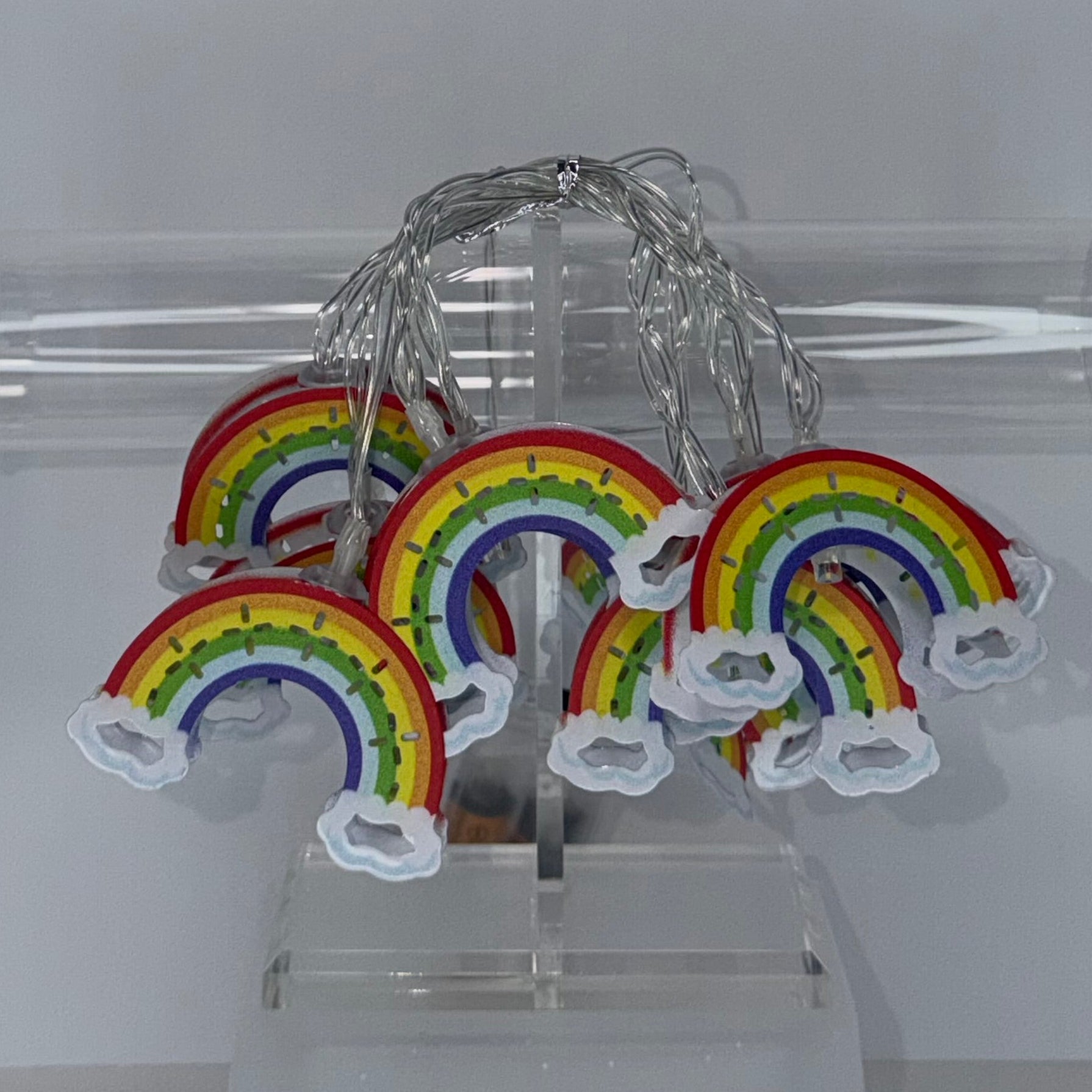 A set of small rainbow-shaped fairy lights with white cloud ends and a silver wire, displayed hanging from a clear acrylic stand in front of a transparent cylindrical tube against a light gray background.