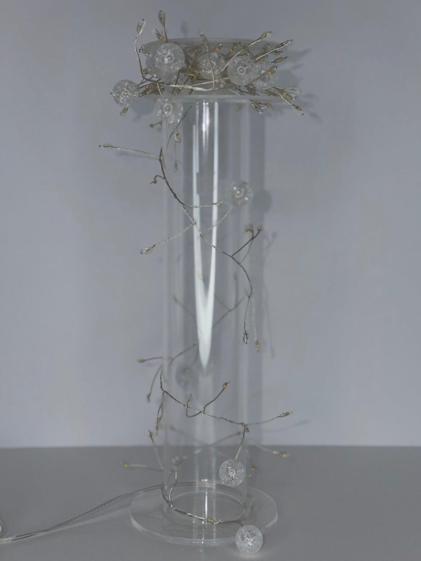 A tall clear cylindrical glass or acrylic pedestal vase with a round base, decorated with delicate silver-toned wire branches and several clear glass orb lights, set against a plain light gray background.