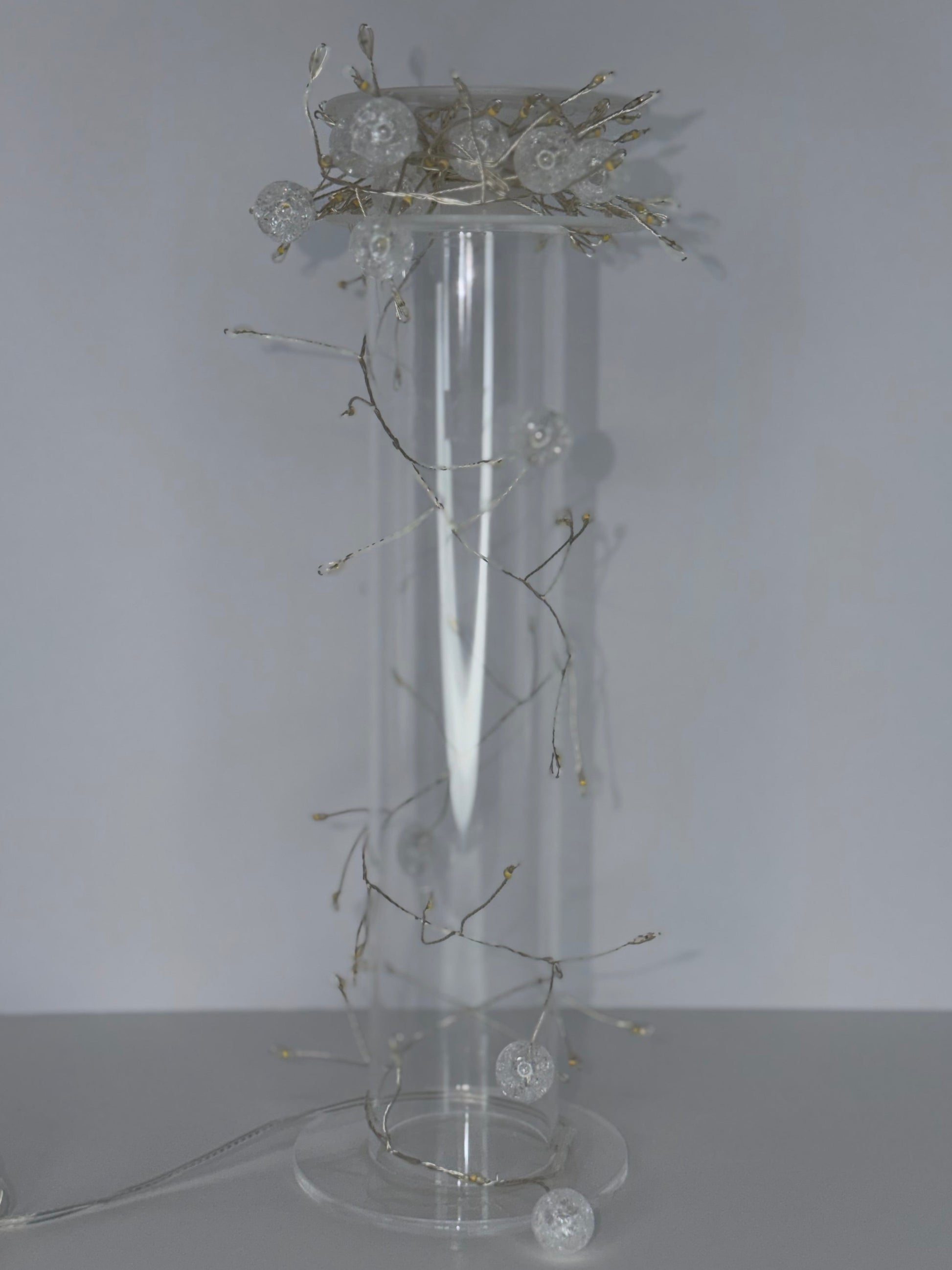 A tall clear cylindrical glass or acrylic pedestal vase with a round base, decorated with delicate silver-toned wire branches and several clear glass orb lights, set against a plain light gray background.