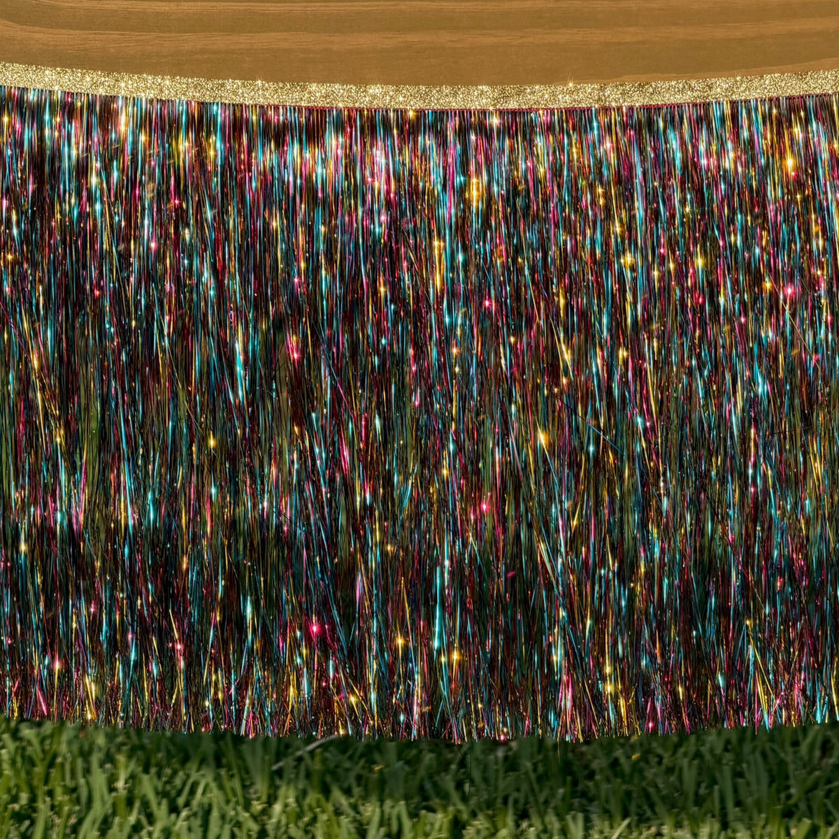 A close-up of a multicolor metallic fringe trim hanging from a wooden surface, with a green grass-like background visible beneath the fringe.