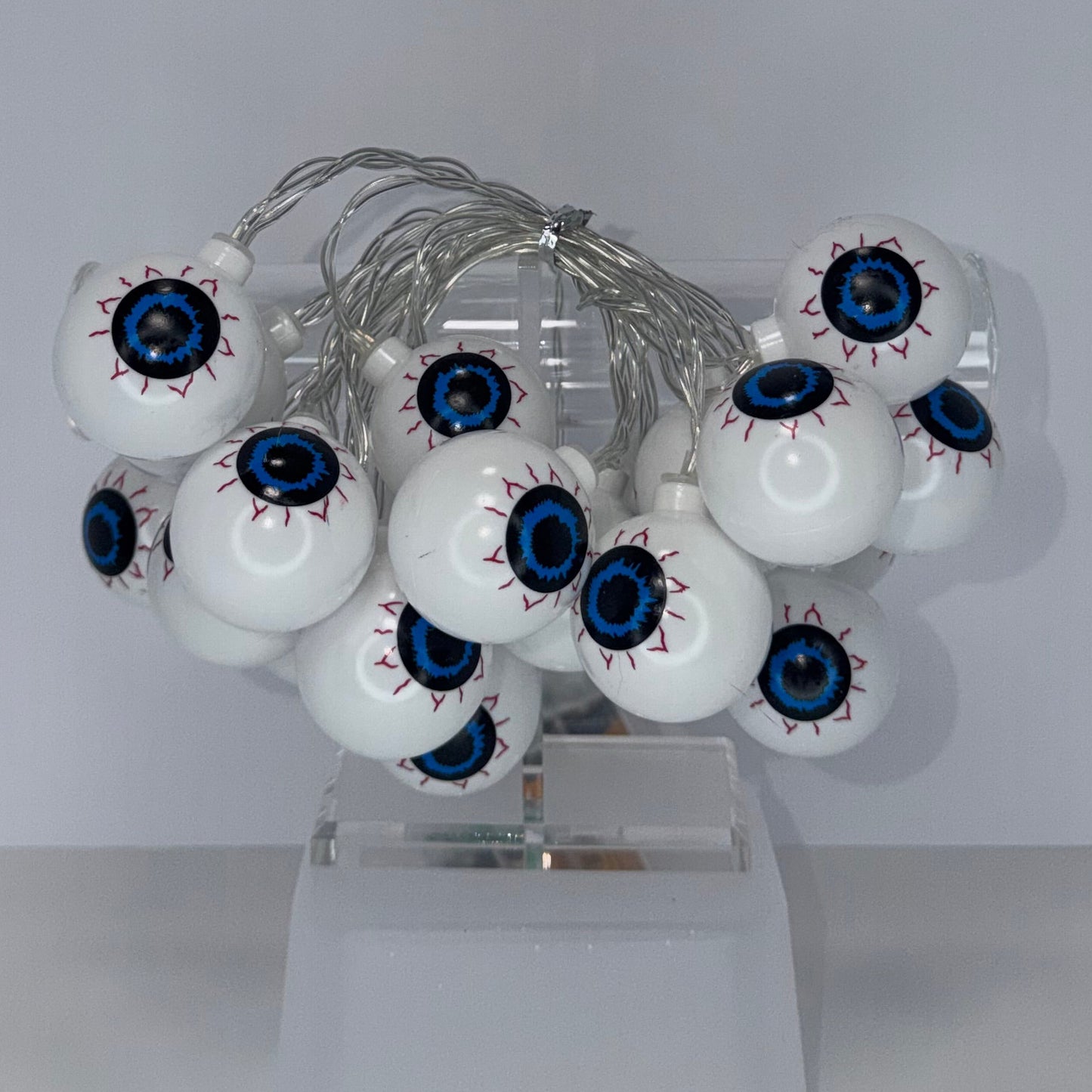 A cluster of white globe-shaped decorative bulbs with detailed blue, black, and red eyeballs, mounted on a clear plastic stand against a plain light gray background.