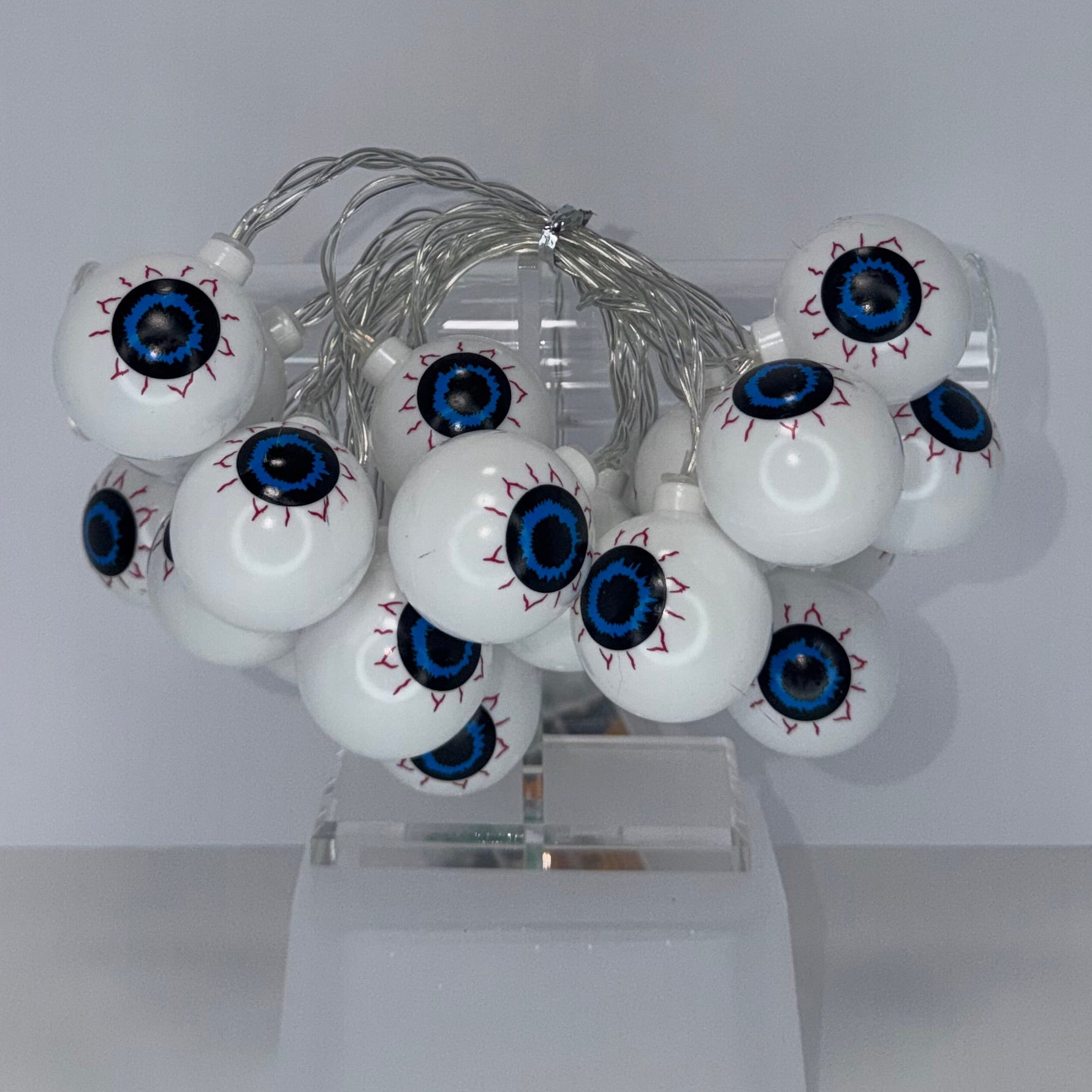 A cluster of white globe-shaped decorative bulbs with detailed blue, black, and red eyeballs, mounted on a clear plastic stand against a plain light gray background.