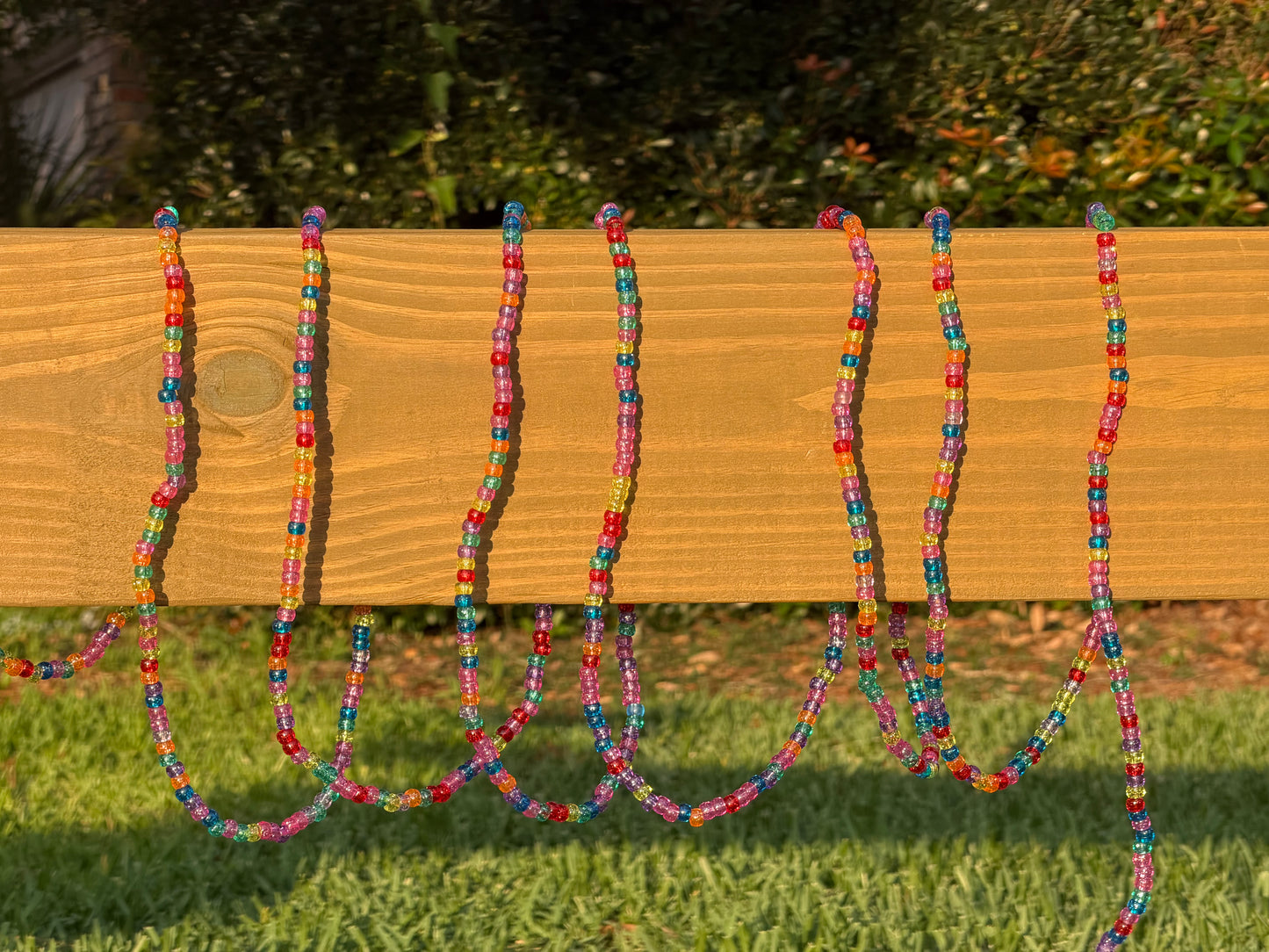 Six colorful beaded garlands draped over a wooden fence outdoors on a sunny day, with green grass and bushes in the background.