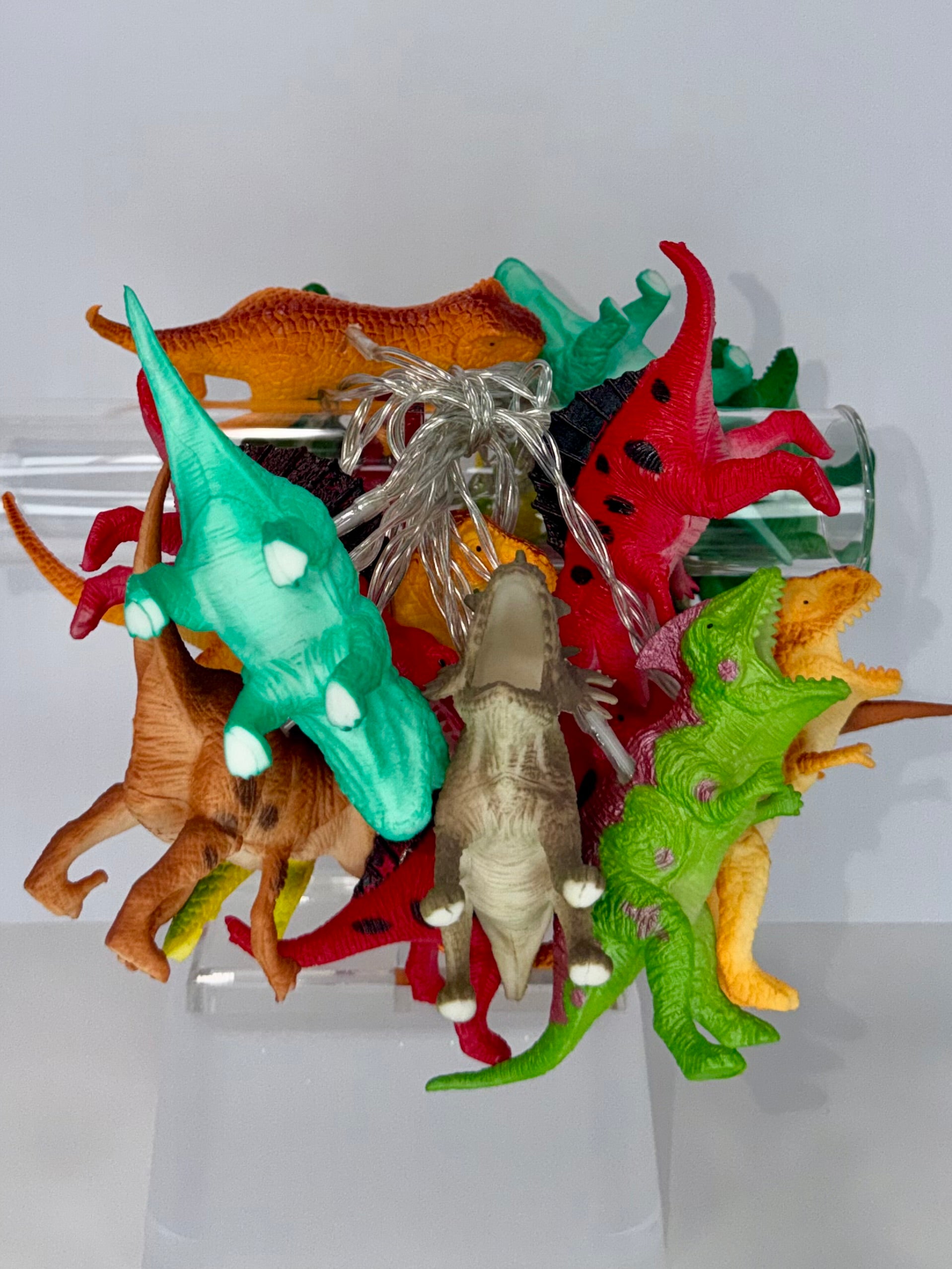 A collection of colorful plastic dinosaur toy figures piled together on a clear acrylic display stand against a plain white background.