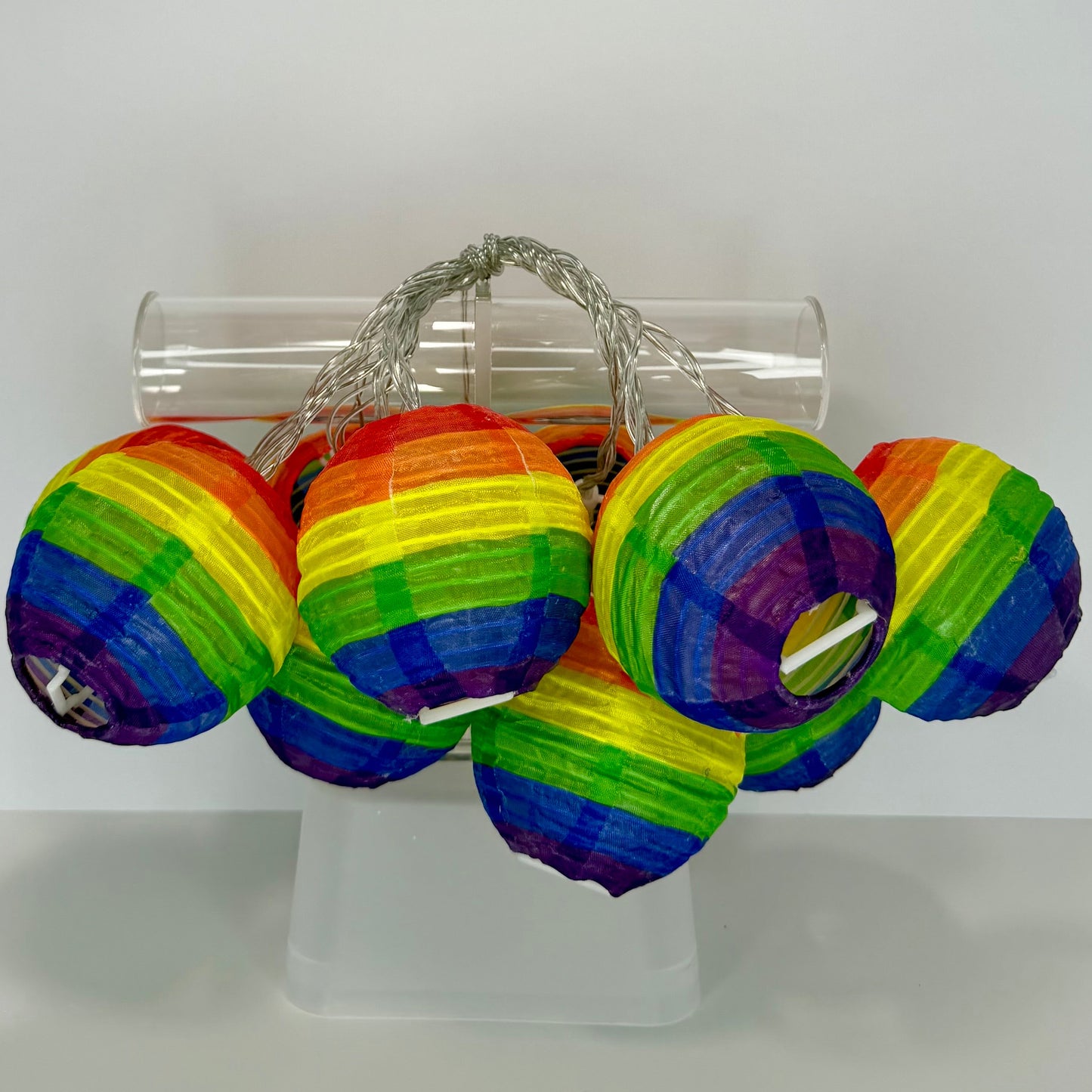A set of small rainbow-striped paper lanterns hanging from a clear plastic tube on a white display stand against a white background.