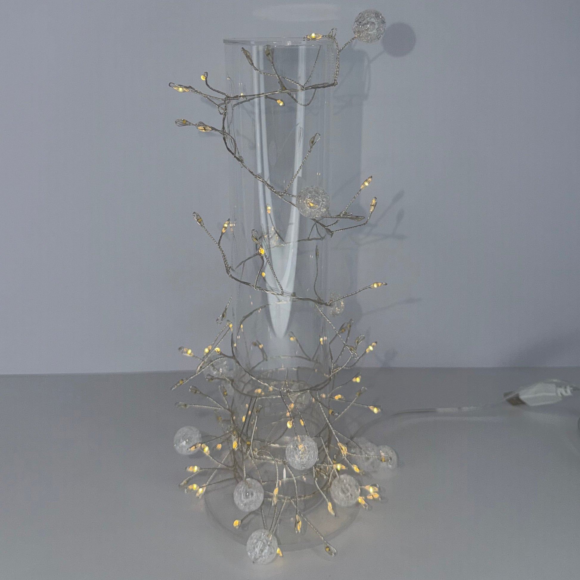 A decorative wall-mounted light fixture with a clear cylindrical glass shade and branching branches covered in warm white fairy lights and white globe bulbs, mounted on a plain light gray wall.