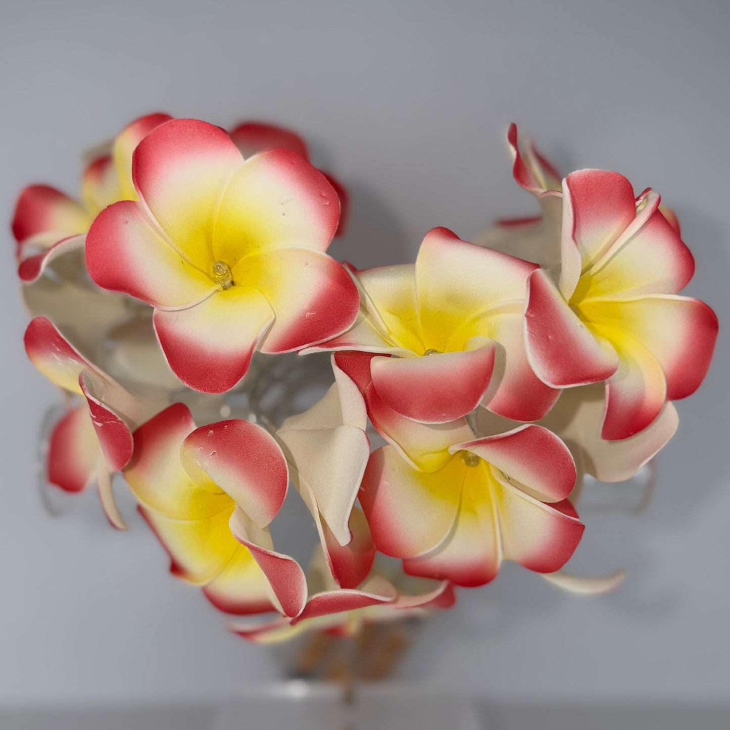 A cluster of artificial plumeria flowers with yellow centers and red-pink edges, arranged on a clear stand against a plain light gray background.
