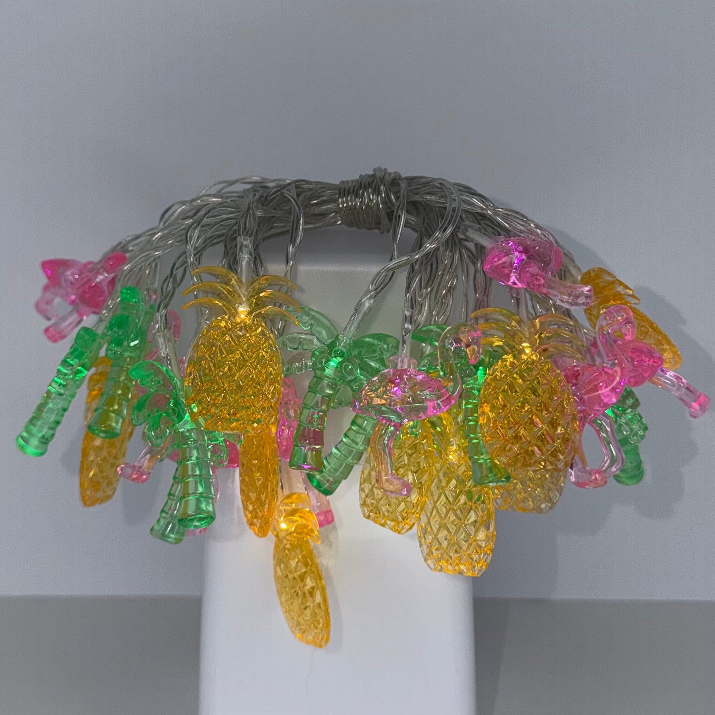 A decorative string light with colorful translucent pineapple-shaped bulbs on a silver wire, mounted on a white wall bracket against a gray wall.