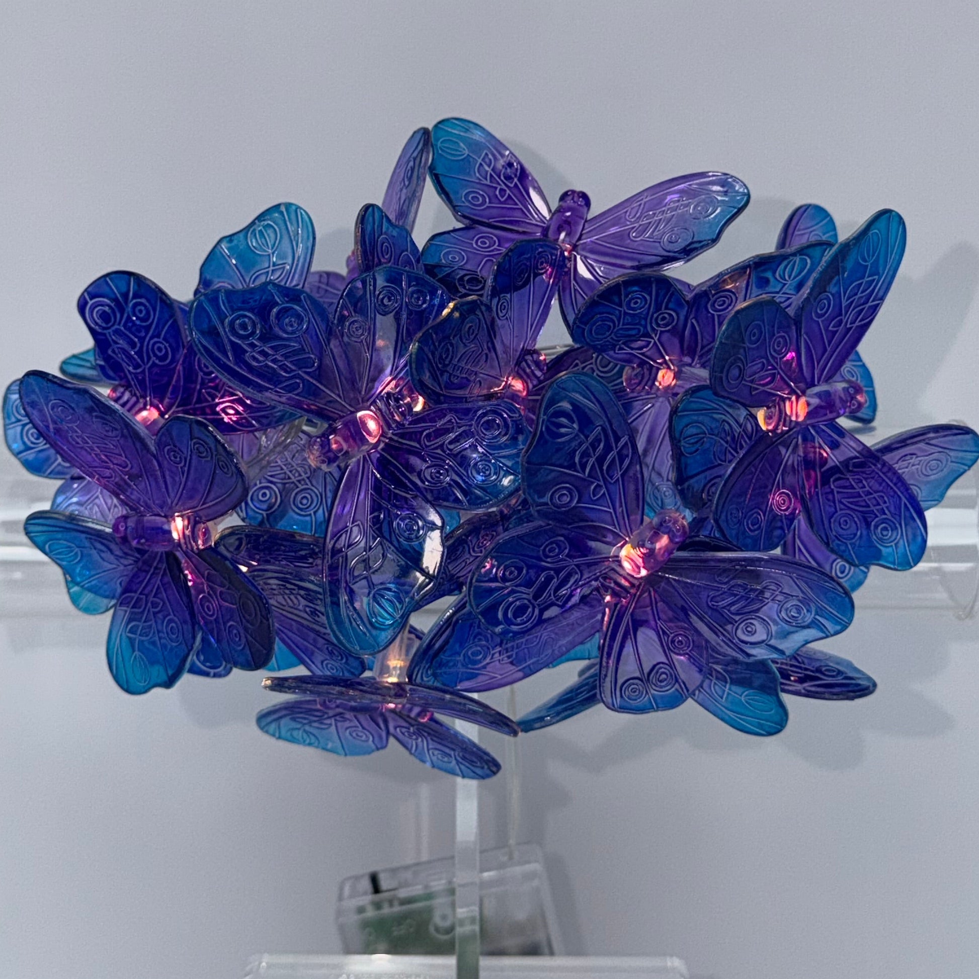 A decorative wall-mounted light fixture shaped like a cluster of blue and purple butterfly wings, with glowing pink lights, mounted on a clear cylindrical base against a plain light gray wall.