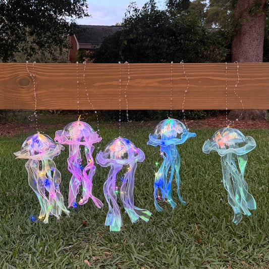 Five hanging jellyfish-shaped solar string lights with iridescent translucent bodies and glowing multicolor accents, displayed outdoors on grass in front of a wooden fence.