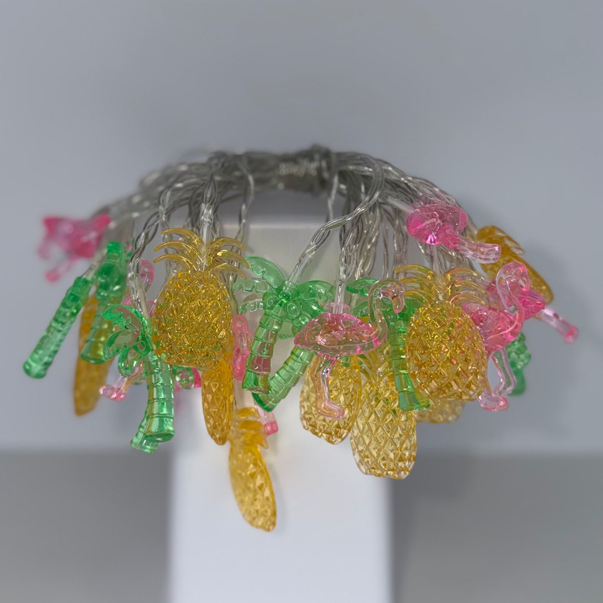A beaded wire crown with yellow pineapple-shaped beads, green leaf-shaped beads, and pink flower-shaped beads on a white display stand.