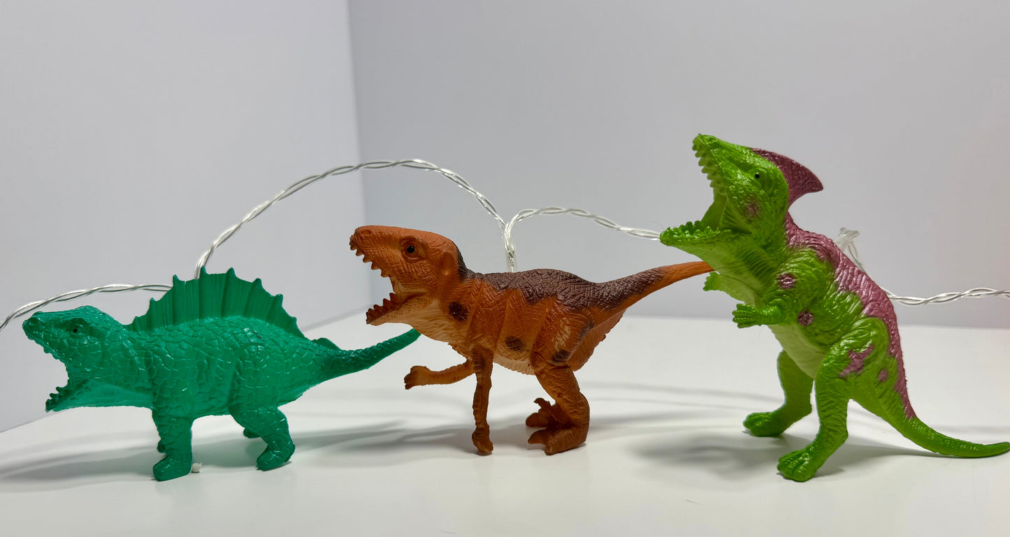 Three small dinosaur figurines arranged on a white surface against a plain light background: a green spiketail on the left, a brown theropod in the center, and a green dinosaur with pink-purple markings on the right. Thin silver string lights are draped behind them.