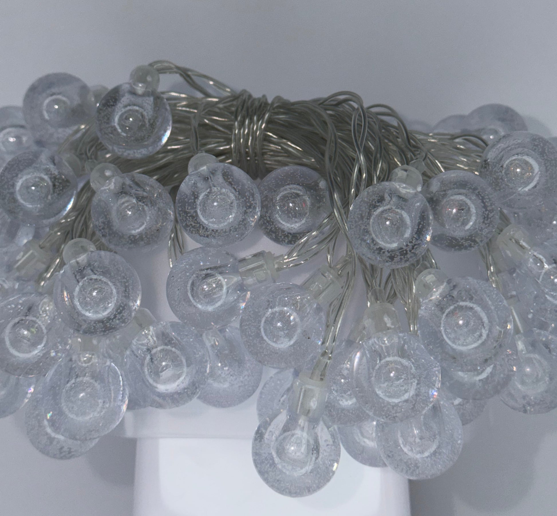 A close-up of a bundle of clear plastic flower-shaped string lights with a silver wire, shown against a plain light gray background.