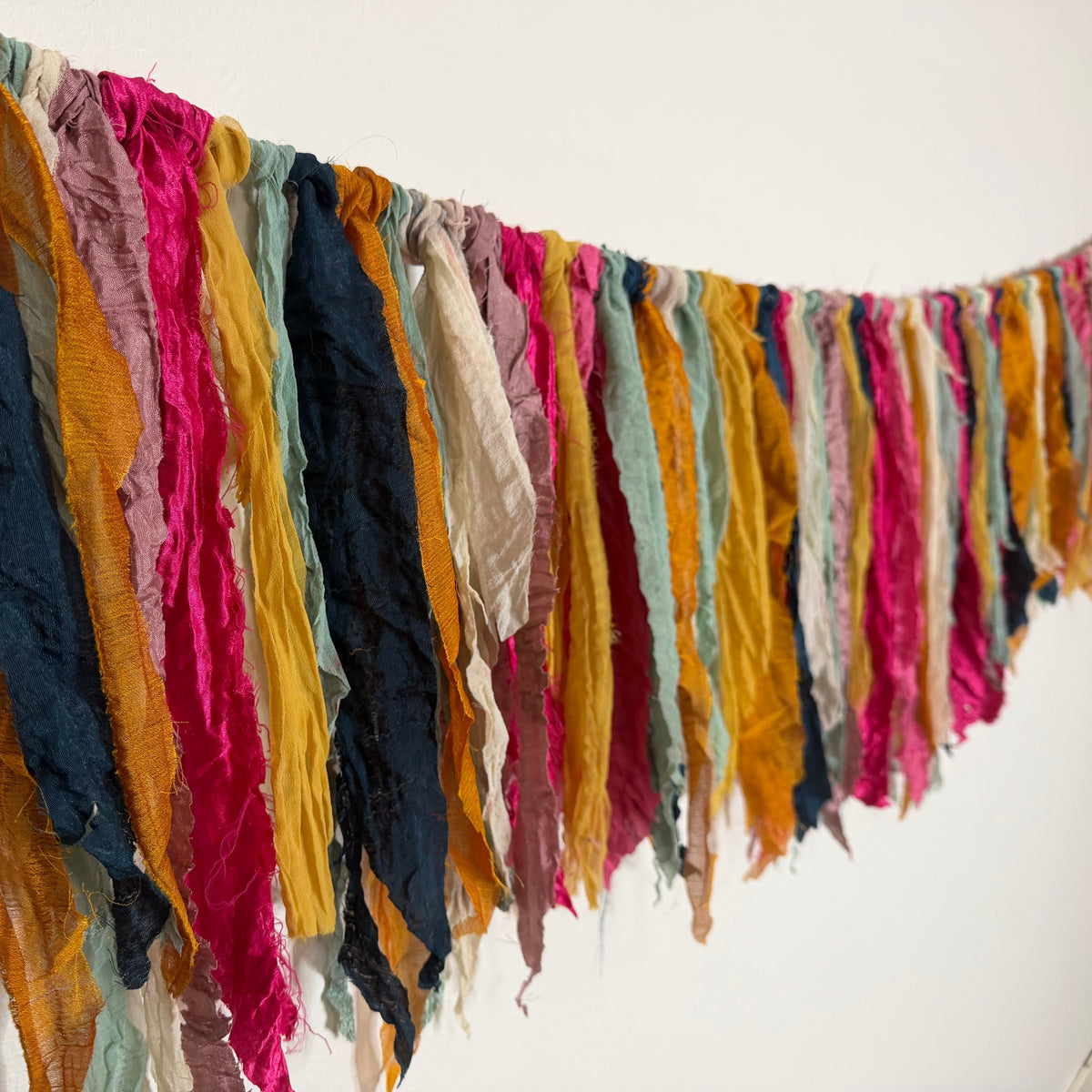 A colorful fabric garland made of hanging crumpled ribbon strips in mustard yellow, pink, navy blue, mint green, and cream, strung along a black cord against a white wall.