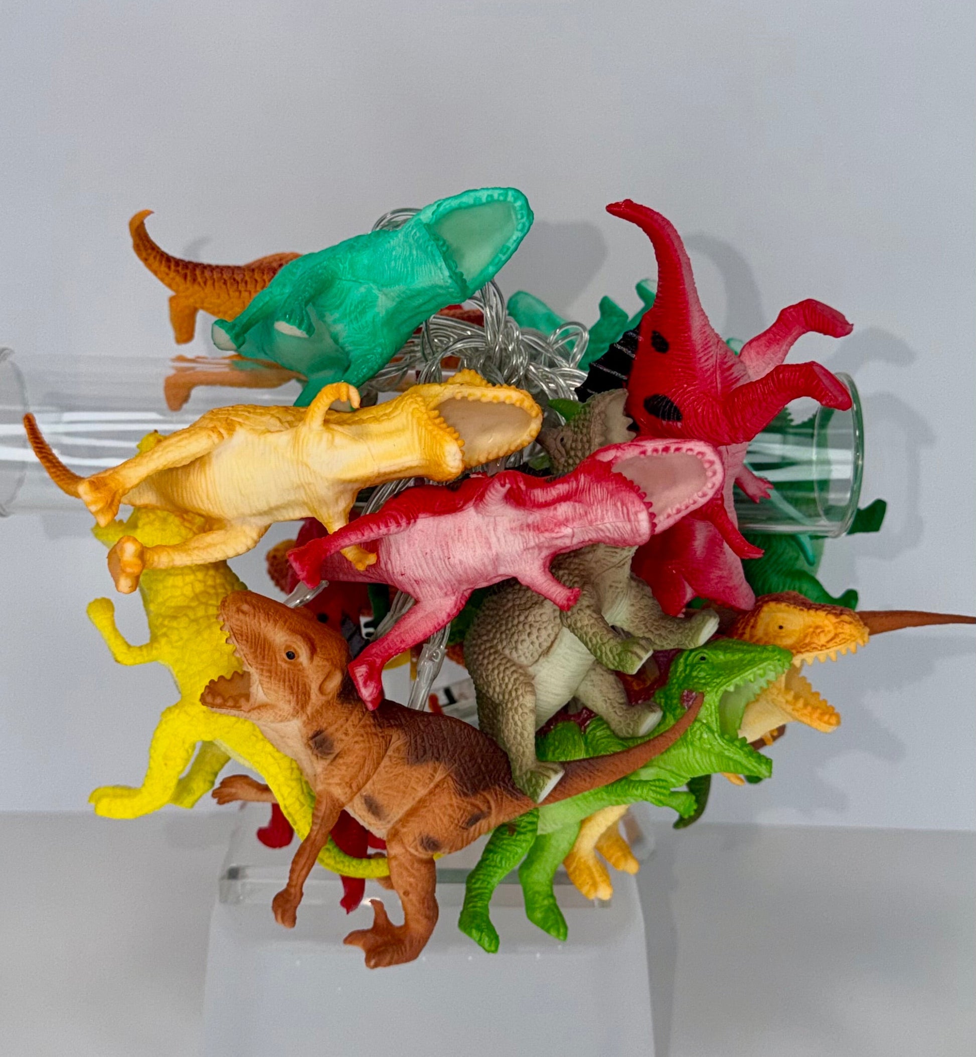 A collection of colorful plastic dinosaur toy figures in various poses, displayed on a clear plastic stand against a light gray background.