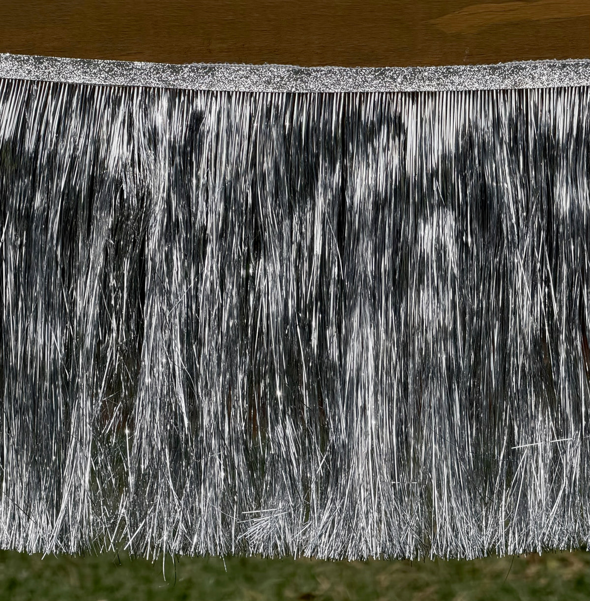 Silver tinsel fringe trim hanging from a glittery silver top band, shown against a background with a wooden surface above and grass below.